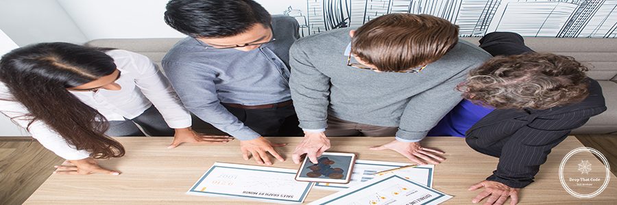 Closeup of four business colleagues discussing diagrams on tablet and papers. They are standing at table. Overhead view.