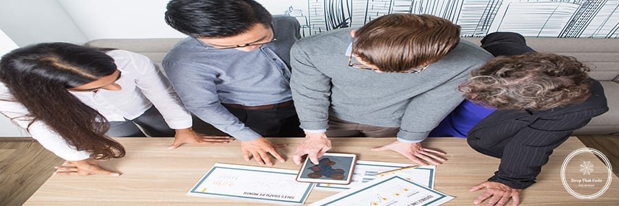Closeup of four business colleagues discussing diagrams on tablet and papers. They are standing at table. Overhead view.