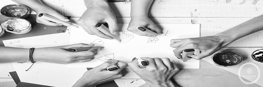 Hands hold colorful markers and draw, top view. Artists wooden table with paints and colored paper. Art and idea concept. Markers in male and female hands draw on white paper with drawings