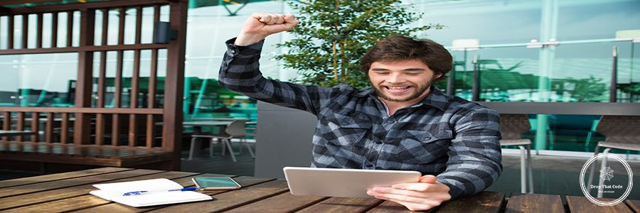 Happy man using tablet and celebrating achievement in cafe. Attractive guy raising clenched hand and sitting at wooden table with outdoor cafe interior in background. Success concept. Front view.