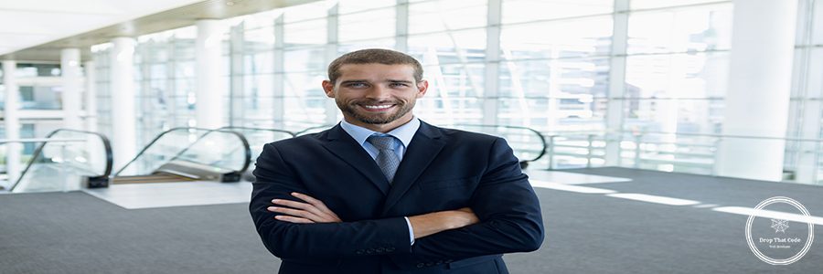 Front view of portrait of young Caucasian businessman with arms crossed smiling to camera. He is standing in the glass walled lobby of a modern business building. Modern corporate start up new business concept with entrepreneur working hard