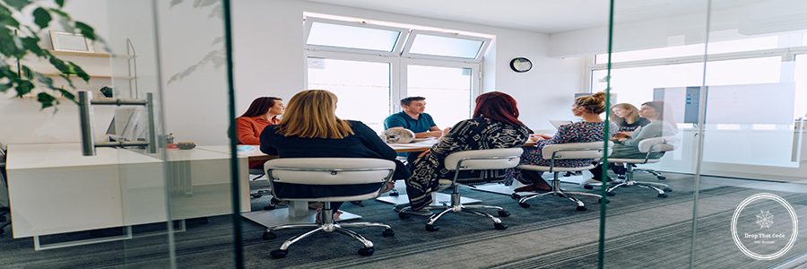 A diverse team of business professionals engaged in a discussion about projects in a modern glass walled office.