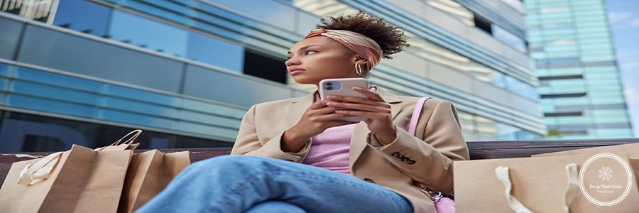 Outdoor shot of thoughtful young African American woman looks pensively away uses mobile phone for surfing social networks poses on bench with many paper shopping bags around enjoys leisure time