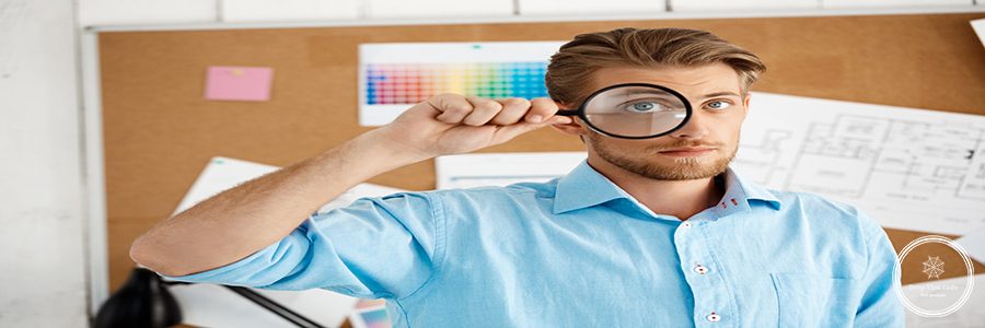 Picture of young handsome confident pensive businessman standing over cork board looking in camera through magnifier. White modern office interior background.